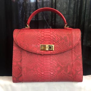 May include: A red handbag with a top handle and a flap closure. The bag has a textured, snakeskin-like pattern and a gold-coloured clasp. The handle is also red and attached with gold-coloured hardware.