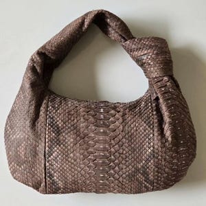 Pode incluir: Uma bolsa marrom com textura de pele de cobra e uma alça com nós. A bolsa tem formato arredondado e uma única alça curva. O padrão de pele de cobra é visível em toda a superfície da bolsa.