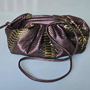 Può includere: Una pochette con motivo a pelle di serpente nei toni del marrone e dell'oro. La pochette ha una chiusura con coulisse e una tracolla intrecciata. La borsa è realizzata in un materiale lucido e ha un design arricciato.