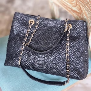 K&ouml;nnte beinhalten: Schwarze, gesteppte Leder-Tote-Tasche mit goldenen Kettenriemen. Die Tasche hat ein Rautenmuster und einen oberen Griff. Die Kettenriemen sind mit goldfarbener Hardware an der Tasche befestigt. Die Tasche liegt auf einer blauen Oberfl&auml;che.