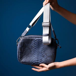 Könnte beinhalten: Eine blaue Umhängetasche mit Schlangenmuster. Die Tasche hat einen verstellbaren silber-weißen Riemen und silberfarbene Hardware. Die Tasche wird vor einem blauen Hintergrund gehalten.