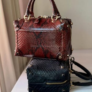 Può includere: Due borse strutturate impilate con manici superiori. La borsa superiore è rossa con un motivo a pelle di serpente, finiture dorate e chiusura con cerniera. La borsa inferiore è nera con un motivo simile, finiture dorate e chiusura con cerniera.