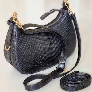 Op de afbeelding: Zwarte, halvemaanvormige handtas met een textuur die op slangenleer lijkt. De tas heeft een goudkleurige rits en hardware, inclusief een afneembare schouderriem. De tas kan over de schouder of als clutch worden gedragen.