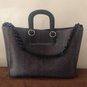 May include: A grey tote bag with a snakeskin pattern. The bag has a top handle and a chain strap. The handle and strap are dark blue. The bag has two silver rings where the strap attaches. The bag is sitting on a brown surface.