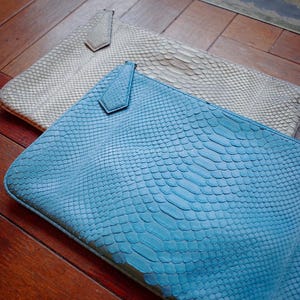 Op de afbeelding: Twee leren clutches met een slangenleerpatroon. Eén is lichtblauw, de andere is gebroken wit. Beide hebben een klein lipje aan de bovenkant. De clutches liggen op een houten oppervlak.