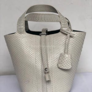 May include: A light grey handbag with a textured snakeskin pattern. The bag has a bucket shape with a top handle and a detachable tag. A strap with a small lock closure is visible. The interior is black.