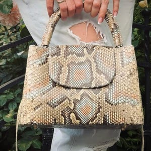 Puede incluir: Un bolso rectangular con cierre de solapa, elaborado con un material similar a la piel de serpiente en tonos beige, marrón y gris. El bolso cuenta con un asa superior y una correa de hombro desmontable. El bolso es sostenido por una persona.