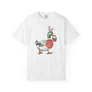 May include: White t-shirt featuring a cartoon duck wearing bunny ears and carrying a basket of eggs. The duck has green, red, and white feathers. The t-shirt is a casual style.