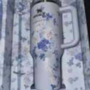 K&ouml;nnte beinhalten: Ein wei&szlig;er Stanley-Becher mit einem floralen Muster in Blau und Gold, mit Griff und transparentem Deckel. Der Becher wird in einer passenden Schachtel mit Blumenmuster zusammen mit einem silbernen Strohhalm pr&auml;sentiert.