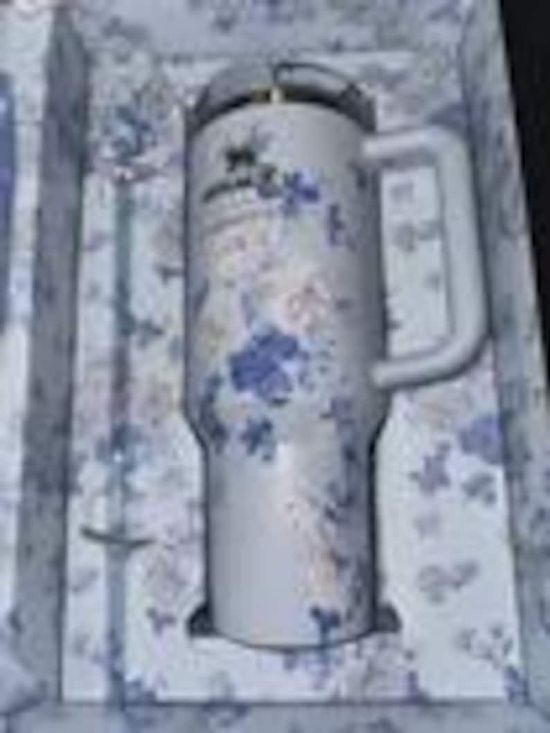 K&ouml;nnte beinhalten: Ein wei&szlig;er Stanley-Becher mit einem floralen Muster in Blau und Gold, mit Griff und transparentem Deckel. Der Becher wird in einer passenden Schachtel mit Blumenmuster zusammen mit einem silbernen Strohhalm pr&auml;sentiert.