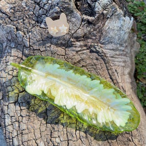 Puede incluir: Una bandeja de resina en forma de hoja con detalles verdes y blancos, y un par de pendientes de gato con detalles de estrella. Los artículos se muestran sobre una superficie de madera envejecida y texturizada.