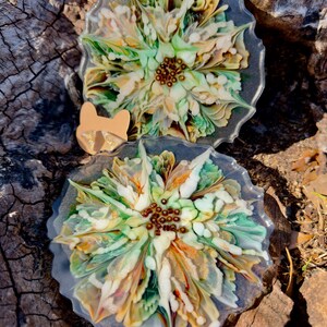 Puede incluir: Dos posavasos de resina con diseños florales. Los posavasos presentan una mezcla de colores verdes, amarillos y blancos, con un grupo central de cuentas marrones. Un pequeño dije en forma de gato de color beige se coloca entre los posavasos.