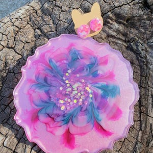 Puede incluir: Un posavasos de resina rosa con un diseño floral en tonos rosa, azul y morado, con detalles de purpurina. Dos pendientes de botón rosas y blancos veteados se muestran sobre un respaldo de cartón con forma de gato.