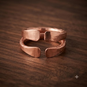 Può includere: Un anello in metallo color rame caratterizzato da un design aperto con due fasce distinte. La superficie del metallo presenta una texture martellata con rientranze visibili e una lucentezza metallica, conferendo un aspetto stratificato al gioiello.