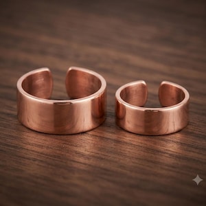Puede incluir: Dos anillos abiertos fabricados en cobre pulido. Los anillos presentan un diseño de banda plana y lisa con un acabado metálico reflectante. Ambas piezas son circulares con una pequeña abertura en la parte posterior, mostrando un aspecto sencillo.