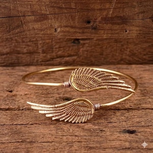 May include: A gold-coloured bracelet featuring two detailed wing designs. The wings are positioned on either side of the bracelet's opening. The bracelet is made of a thin, flexible metal and rests on a wooden surface.