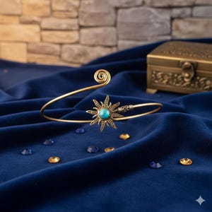 Peut inclure: Un bracelet de bras doré avec un motif de soleil et une pierre centrale turquoise. Le bracelet a un détail en spirale à une extrémité et repose sur une surface de velours bleu foncé. Des pierres précieuses décoratives et une petite boîte sont en arrière-plan.