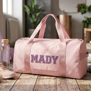 Peut inclure: Un sac de sport rose clair avec le nom MADY en lettres violettes pailletées sur le devant. Le sac possède deux poignées supérieures et une fermeture éclair. À proximité se trouvent un tapis de yoga violet clair, une gourde, des baskets rose clair et des serviettes pliées.