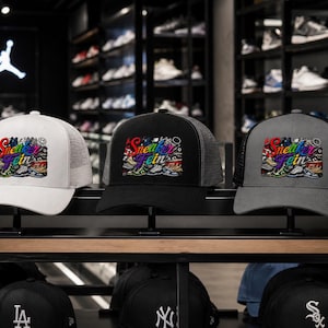 May include: Three trucker hats with the text "Sneaker Fein" in a colorful design. The hats are white, black, and gray, with mesh backs. The hats are displayed on a shelf in a shoe store, with many pairs of sneakers in the background.