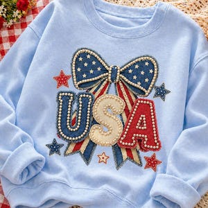 May include: Pale blue sweatshirt featuring a patriotic USA design. The design includes a denim bow with white stars, red and white stripes, and the letters "USA" with a pearl trim. Red and blue stars complete the design.