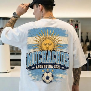 May include: A white short-sleeve t-shirt featuring a distressed graphic on the back. The design includes a yellow sun with a face, the text "MUCHACHOS ARGENTINA 2026" in blue, and a soccer ball graphic over a light blue and white striped background.