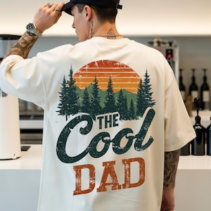 May include: An off-white short-sleeve t-shirt featuring a graphic on the back. The design displays a forest of green pine trees under an orange sun, with the text "THE Cool DAD" printed in dark green and orange with a distressed texture.