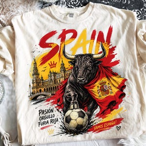 May include: A cream-colored t-shirt featuring a graphic of a bull draped in the Spanish flag, standing over a soccer ball. The design includes the text SPAIN, PASIÓN ORGULLO FURIA ROJA, and VAMOS ESPAÑA, alongside an illustration of a building.