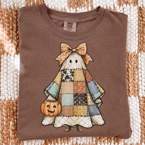 May include: A brown t-shirt featuring a graphic of a ghost wearing a patchwork quilt costume with a bow on its head and holding a jack-o'-lantern bucket. The shirt has a crew neckline and a Comfort Colors brand label printed on the inside collar.