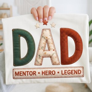 Könnte beinhalten: Cremefarbener Stoff mit dem Wort "DAD" in grünen, floralen und Cordbuchstaben. Das Wort "DAD" steht über den Wörtern "MENTOR • HERO • LEGEND" in einem rechteckigen Rahmen. Drei sternförmige Verzierungen befinden sich über dem Wort "DAD".