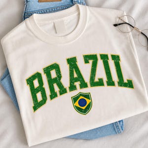 May include: A white short-sleeved t-shirt featuring the word BRAZIL in large, distressed green block letters with a yellow outline. Below the text is a small shield-shaped emblem containing a yellow diamond, a blue circle, and green borders.