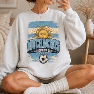 May include: A light gray crewneck sweatshirt featuring a distressed graphic of the Argentine flag with a central sun emblem. The text reads "MUCHACHOS" and "ARGENTINA 2026" above a soccer ball graphic with three small stars arranged in an arc above it.
