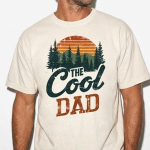 May include: A cream-colored short-sleeve t-shirt featuring a graphic print of a sunset over a forest. The design includes the text "THE COOL DAD" in a distressed font, with "THE COOL" in dark green script and "DAD" in a bold, blocky brown font.