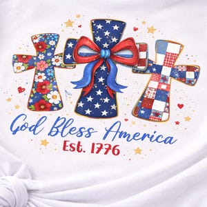 Può includere: T-shirt bianco con un disegno di tre croci decorative con motivi patriottici. La croce centrale ha un fiocco rosso, bianco e blu. Il testo "God Bless America Est. 1776" è stampato sotto.