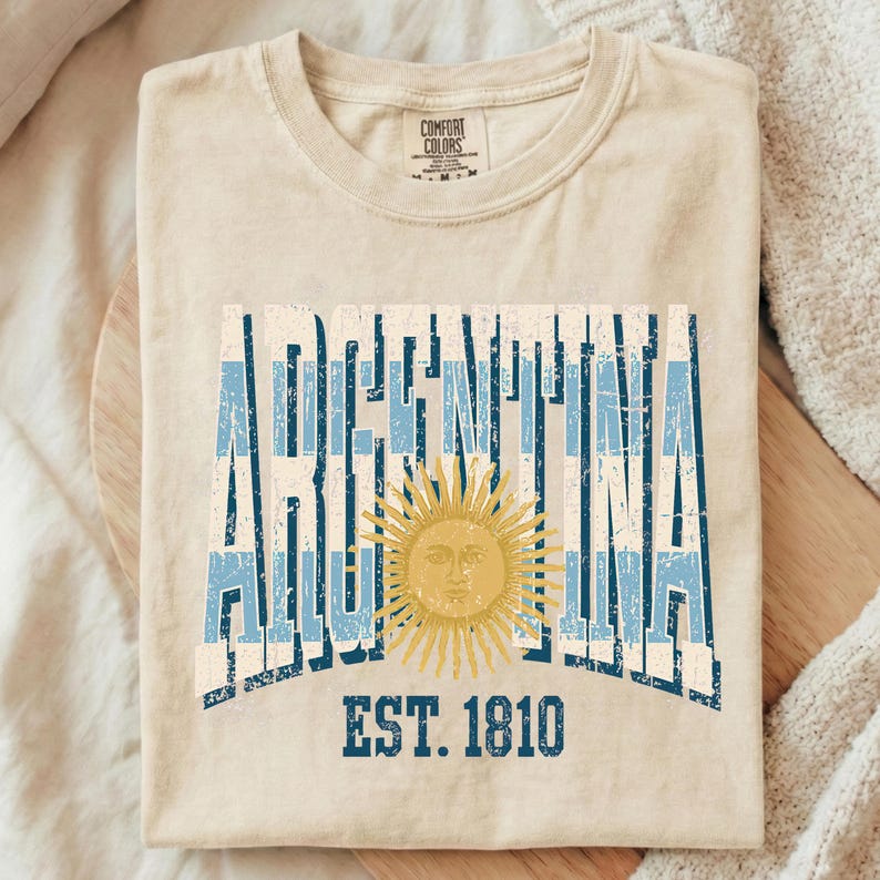 May include: A beige crew neck t-shirt featuring the word ARGENTINA in a distressed blue and white striped font. A golden yellow sun with a human face is centered over the text. Below the sun, the text EST. 1810 is printed in blue.
