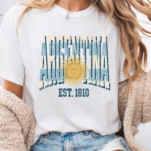 May include: A white t-shirt featuring a distressed graphic design with the word ARGENTINA in dark blue block letters over light blue vertical stripes. A yellow sun with a face is centered in the design, with the text EST. 1810 printed below.