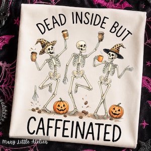 Puede incluir: Una camiseta blanca con un gr&aacute;fico de tres esqueletos bailando con tazas de caf&eacute; y calabazas. Dos esqueletos llevan sombreros de bruja con estampado de leopardo. El texto DEAD INSIDE BUT CAFFEINATED aparece escrito en letras negras.