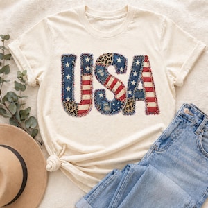 May include: Cream-colored t-shirt with the word "USA" in a patchwork design. The letters feature denim, leopard print, and red and white stripes, evoking the American flag. A pair of blue jeans and a hat are visible.
