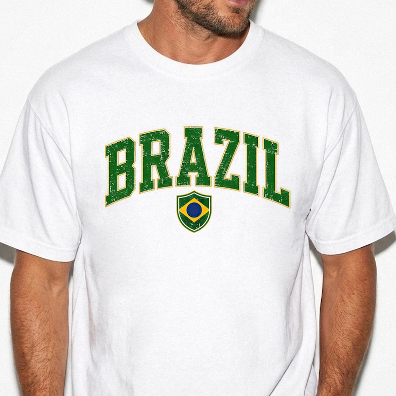 May include: A white short-sleeved t-shirt featuring the word BRAZIL printed in a distressed green font with a yellow outline in an arched layout. Below the text is a small shield-shaped emblem containing a yellow diamond, a blue circle, and a green border.