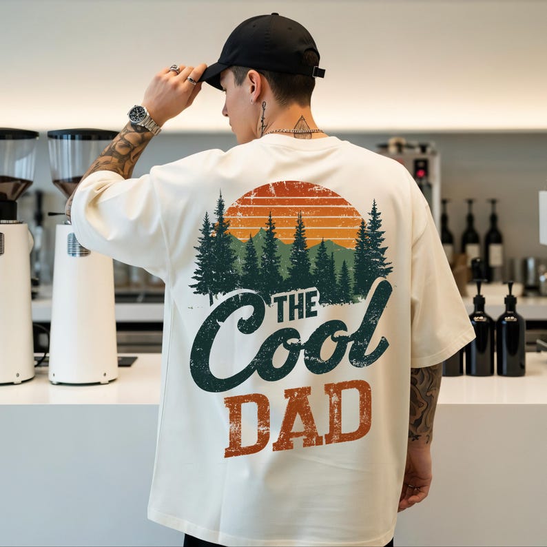 May include: An off-white short-sleeve t-shirt featuring a graphic on the back. The design displays a forest of green pine trees under an orange sun, with the text "THE Cool DAD" printed in dark green and orange with a distressed texture.