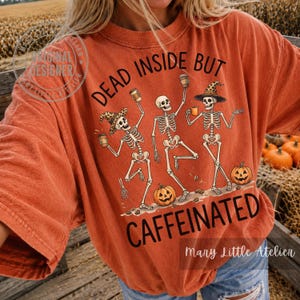 Puede incluir: Una camiseta naranja con un gr&aacute;fico de tres esqueletos bailando con tazas de caf&eacute; y calabazas. El texto dice "DEAD INSIDE BUT CAFFEINATED". Los esqueletos llevan sombreros y el dise&ntilde;o incluye granos de caf&eacute; dispersos y calabazas naranjas.