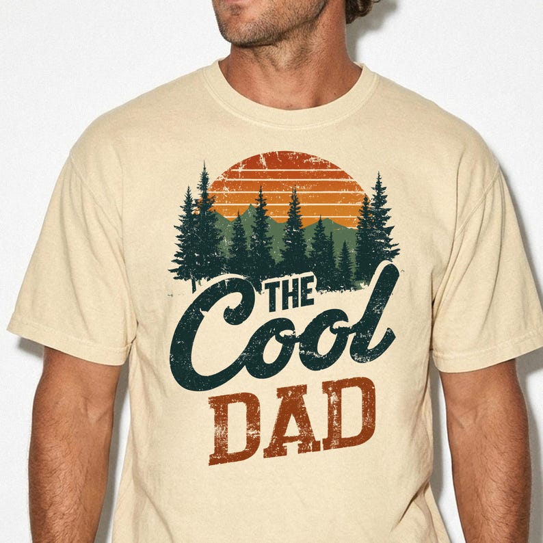 May include: A beige short-sleeve t-shirt featuring a graphic print of a sunset over a forest of pine trees. The text "THE COOL DAD" is printed in a distressed font below the trees, with "THE COOL" in dark blue and "DAD" in a rust orange color.