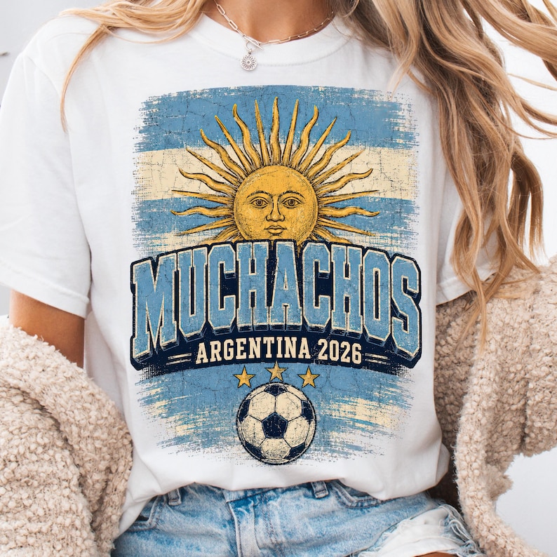 May include: A white t-shirt featuring a distressed graphic of the Argentine flag with a central sun emblem. The text reads MUCHACHOS ARGENTINA 2026 in bold blue letters above a soccer ball illustration with three small yellow stars positioned above it.