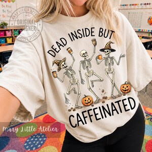Puede incluir: Una camiseta color crema con un gr&aacute;fico de tres esqueletos bailando que sostienen tazas de caf&eacute;. Dos esqueletos llevan sombreros con estampado de leopardo. Hay dos calabazas naranjas en la parte inferior. El texto dice: DEAD INSIDE BUT CAFFEINATED.