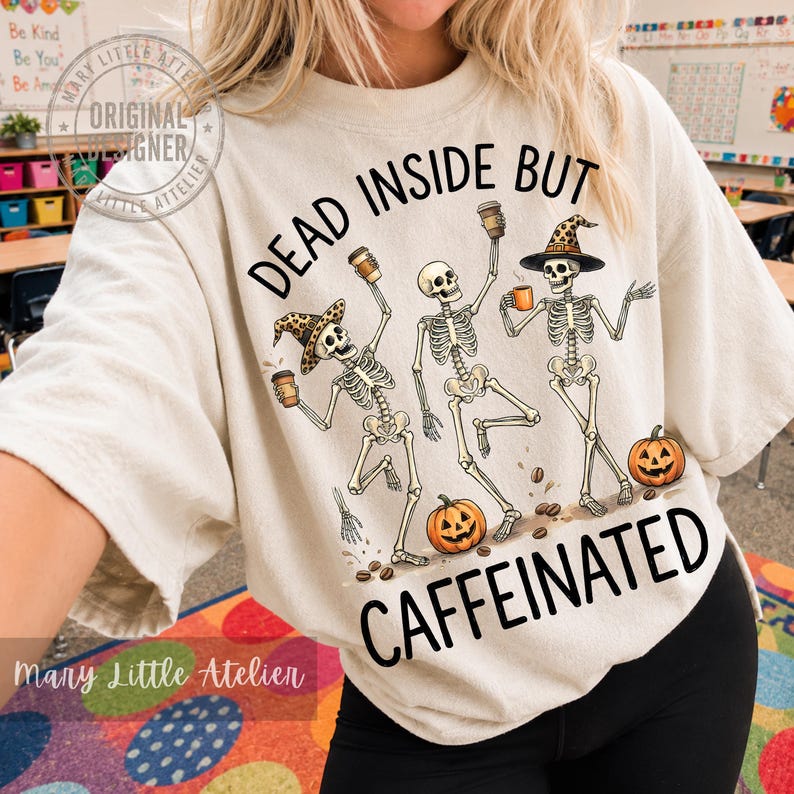 Puede incluir: Una camiseta color crema con un gr&aacute;fico de tres esqueletos bailando que sostienen tazas de caf&eacute;. Dos esqueletos llevan sombreros con estampado de leopardo. Hay dos calabazas naranjas en la parte inferior. El texto dice: DEAD INSIDE BUT CAFFEINATED.