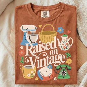 May include: A rust-colored t-shirt featuring a vintage-themed graphic. The design includes a stand mixer, basket, teapot, teacup, purse, and a rotary phone. The words "Raised on Vintage" are prominently displayed in a retro font, surrounded by floral accents.