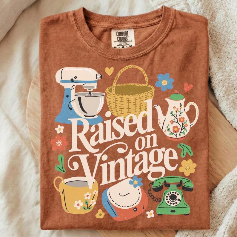 May include: A rust-colored t-shirt featuring a vintage-themed graphic. The design includes a stand mixer, basket, teapot, teacup, purse, and a rotary phone. The words "Raised on Vintage" are prominently displayed in a retro font, surrounded by floral accents.