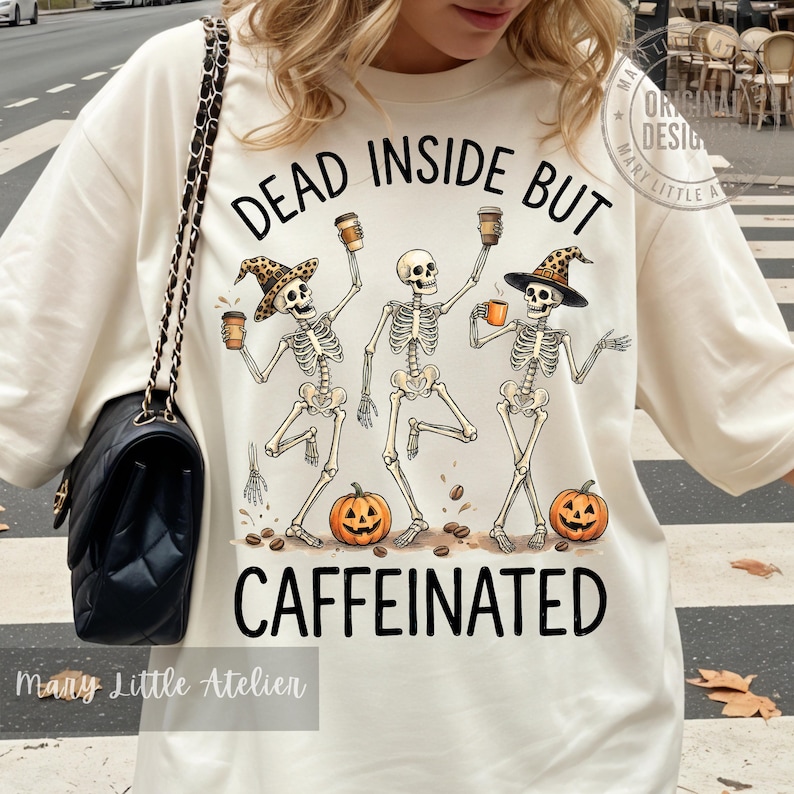 Puede incluir: Una camiseta de color crema con un gr&aacute;fico de tres esqueletos que sostienen tazas de caf&eacute; y llevan sombreros, junto a dos calabazas y granos de caf&eacute;. El texto "DEAD INSIDE BUT CAFFEINATED" aparece en letras negras sobre y bajo los esqueletos.