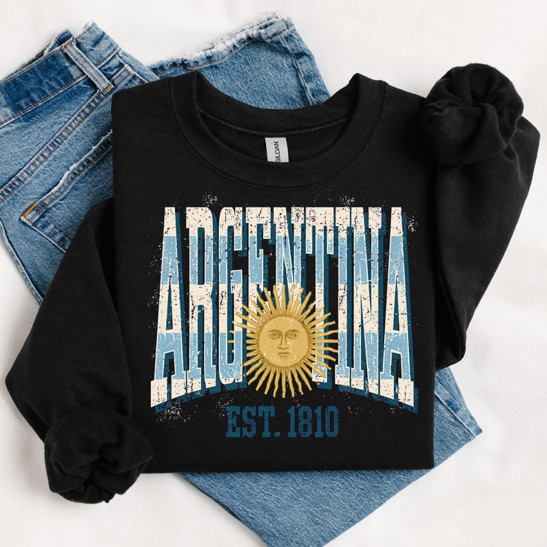 May include: A black long-sleeve sweatshirt featuring the word ARGENTINA in large, distressed white and light blue block letters. A yellow sun graphic with a face is centered over the text, with the blue text EST. 1810 printed below it.