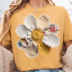 Peut inclure: T-shirt jaune moutarde orné d'une grande fleur appliquée. La fleur a un centre jaune et des pétales faits de divers tissus à motifs, dont du vichy et des imprimés floraux. Les bords des pétales sont festonnés.