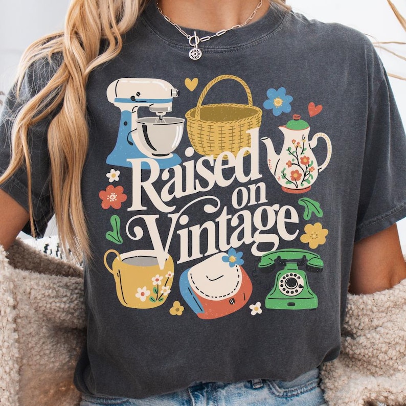 May include: Dark grey t-shirt featuring a colourful graphic design. The design includes a blue stand mixer, a woven basket, a teapot, a teacup, a purse, and a green rotary phone. The words "Raised on Vintage" are in the centre.